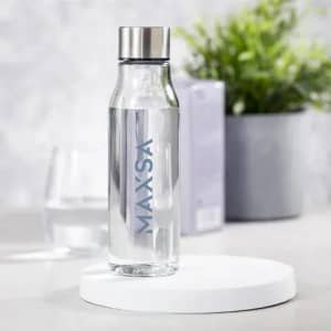 A drink bottle made of clear glass with a stainless steel lid, placed on a white base against a blurred background.
