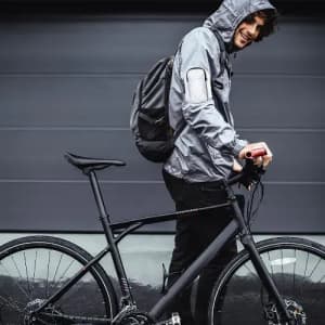 A man in a grey hooded jacket stands next to a black bicycle, holding the handlebars while smiling.