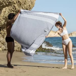A beach scene with a man and woman holding a large grey and white striped towel, set against a rocky shoreline and blue sky.