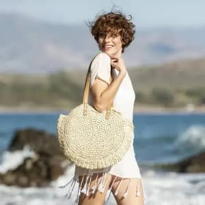 A woven straw beach bag with wooden handles, set against a coastal backdrop with waves and rocks.