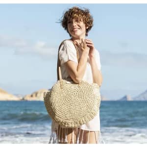 A woven straw bag with a round shape, held by a smiling woman on a beach, with ocean waves in the background.