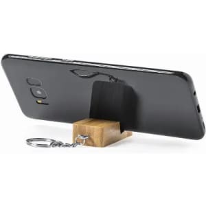 A phone stand displaying a black smartphone propped on a wooden base with a keychain attachment.