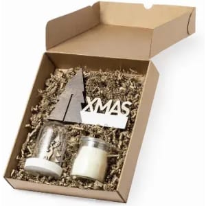 A gift box containing two candles, a wooden tree decoration, and "XMAS" lettering on a bed of shredded paper.