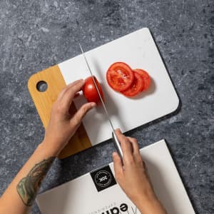A wooden and white chopping board with a hand slicing red tomatoes on it, set against a grey-textured background.