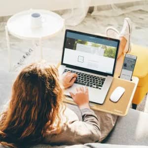 A laptop on a wooden lap desk, with a mouse and a smartphone, beside a cup on a small table. The setting is bright and airy.