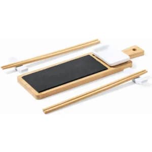 A bamboo sushi serving board featuring a black slate area, with chopsticks and a small white sauce dish.