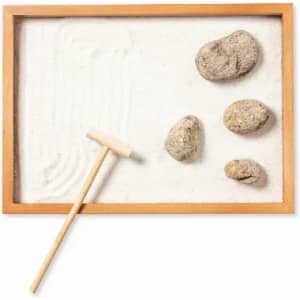 A sand tray with a wooden frame, featuring smooth stones and a small rake. The sand has gentle patterns.