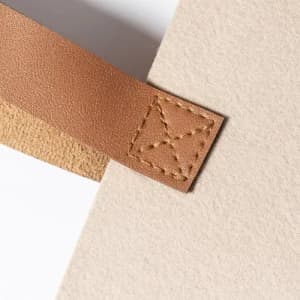 A close-up of a light beige felt product with a brown leather strap and stitched detail.