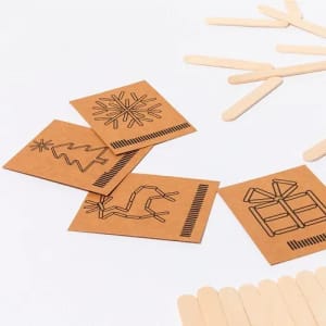 A collection of brown craft cards featuring black line drawings of a snowflake, tree, cactus, and gift, alongside wooden sticks.