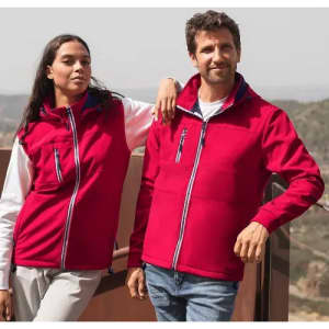 A couple stands together wearing bright red jackets, set against a scenic outdoor backdrop. The jackets feature zippers and pockets.