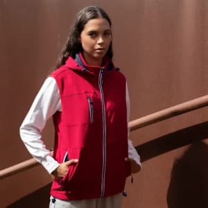 A woman wearing a red sleeveless jacket with a hood stands against a brown wall, hands in pockets.