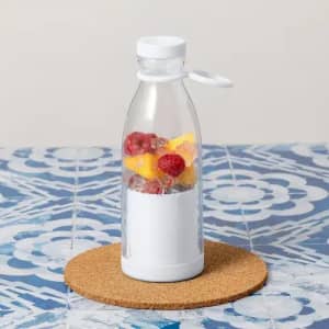 A drink bottle with a white base and clear body contains diced mango and raspberries, sitting on a cork mat.