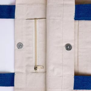 A cream-coloured bag featuring blue handles and a zippered pocket on the inside, along with metal fasteners.