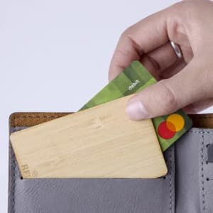 A hand is placing a wooden card and a debit card into a grey wallet, showcasing their contents.