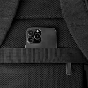 A black backpack featuring a pocket that holds a black smartphone. The material appears durable and textured.