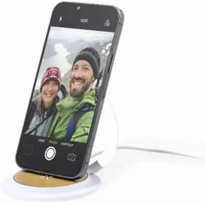 A smartphone on a circular wooden charging pad, displaying a selfie of two people in outdoor attire.