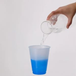 A drink bottle pouring clear liquid into a translucent cup with a blue bottom. The background is plain white.