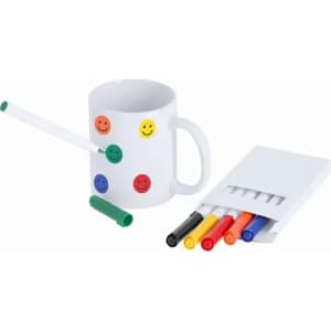 A white mug with smiley face stickers, a green pen, and a set of colourful markers in a case.