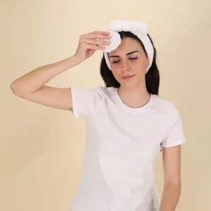A person wearing a white headband holds a cotton pad, appearing to cleanse their face, dressed in a white T-shirt.