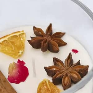 A candle with a white wax base featuring cinnamon, dried orange, star anise, and rose petals.
