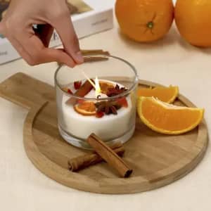 A glass candle with orange slices and cinnamon sticks on a wooden tray, surrounded by fresh oranges.