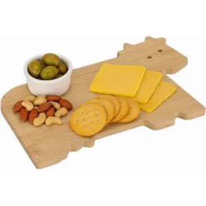 A wooden serving board shaped like a cow, featuring cheese slices, crackers, nuts, and green olives.