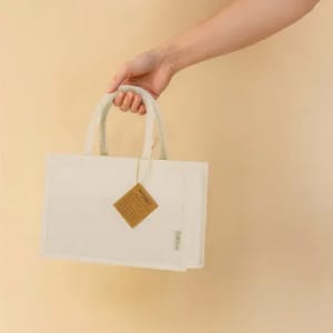 A hand holds a beige, rectangular tote bag with two handles and a tag attached.