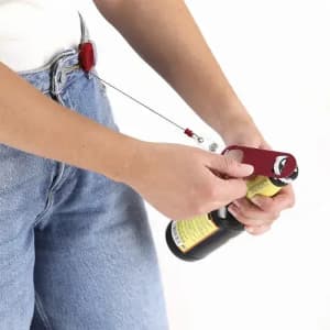 A drink bottle opener with a red handle, attached to a bottle, held by a person wearing denim jeans.