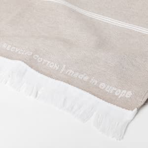A light beige cotton towel with white fringe detailing and text woven into the fabric. It has a soft texture.