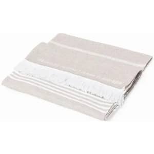 A folded towel in light beige and grey, featuring white fringes and decorative stripes. It has a subtle branding logo.