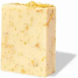 A bar of soap in cream colour with embedded natural herbs and floral elements. The soap has a textured surface.