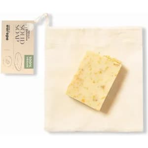 A solid soap bar in a natural cream colour, packaged in a small fabric bag. The soap includes visible exfoliating particles.