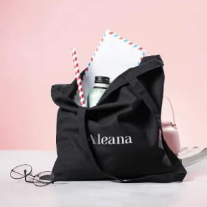 A black tote bag with a logo, containing a drink bottle, papers, and a straw, set against a pink background.
