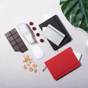 A collection of notebooks in red, black, and silver, with chocolate, raspberries, milk, and granola scattered around.