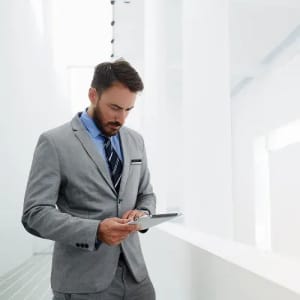 A man in a grey suit and blue shirt stands, looking at a tablet in a modern, bright environment.