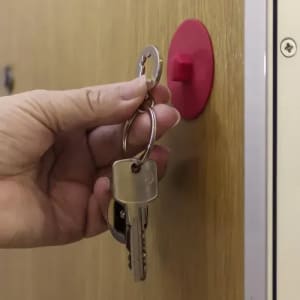 A hand holding a set of keys, noting a red wall-mounted key hook in the background.
