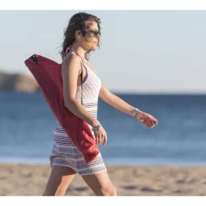 A woman in a striped dress walks along the beach, carrying a red item over her shoulder. The ocean is in the background.