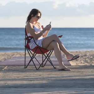A woman sitting on a beach, wearing sunglasses and a striped outfit, using a phone while seated in a red beach chair.