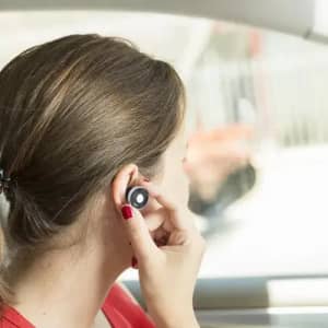 A woman with brown hair is wearing a wireless earbud while seated in a car. The earbud is black.