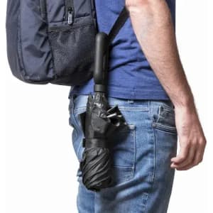 A black umbrella is securely attached to the side of a person’s backpack, worn over blue jeans.