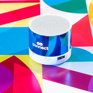 A speaker with a shiny blue finish and a white base, featuring a logo and positioned on a brightly coloured geometric surface.