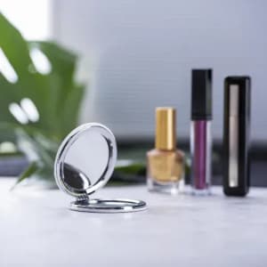 A compact mirror with a shiny silver finish, surrounded by nail polish and lip gloss in various colours.