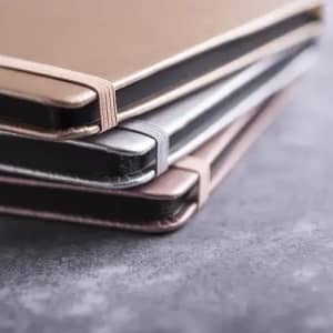 A stack of three notebooks in gold, silver, and black with elastic closures.
