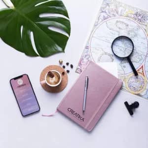 A pink notebook with a pen, a cup of coffee, a phone, a magnifying glass, and a leafy plant on a flat surface.