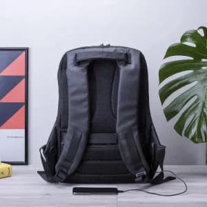 A black backpack with padded straps and a USB charging port, displayed against a neutral background with a plant.