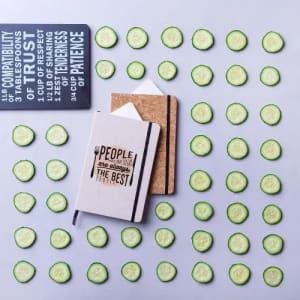 A notebook lies on a surface surrounded by slices of cucumber, with a second notebook partially visible nearby.