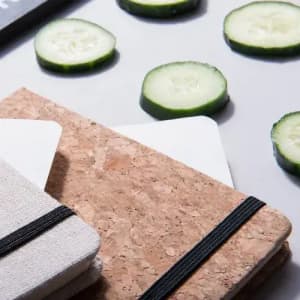 A collection of notebooks with cork and fabric covers, surrounded by sliced cucumber on a flat surface.