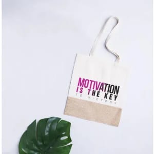 A tote bag in cream and jute with a graphic design featuring the words "Motivation is the Key to Victory."
