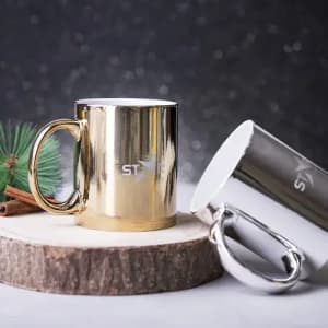 A gold and a silver mug are positioned on a wooden surface, featuring a logo on each. The background is softly lit.