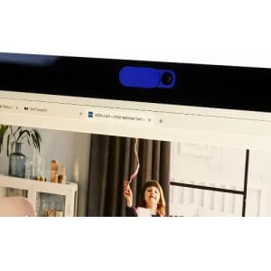 A webcam cover in blue positioned on a computer monitor. The background features a home interior with a child playing.